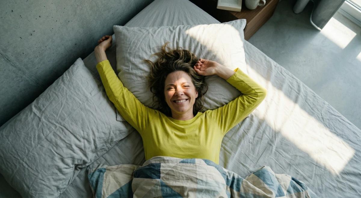 Woman in a yellow top lying on a bed with arms stretched overhead, smiling up at the camera in a bright room, embracing a positive morning routine for well-being