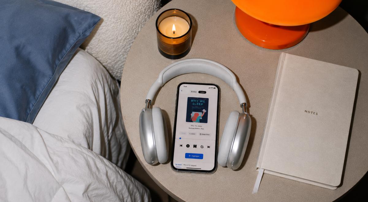 Smartphone playing the Why We Sleep audiobook, surrounded by silver headphones on a bedside table with a lit candle, notebook and orange lamp, ideal for audiobooks for sleep