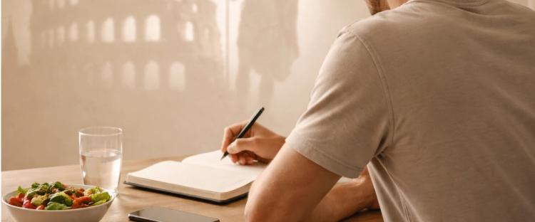 Man writing in a notebook at a wooden desk with a salad, water glass, phone, and earbuds nearby, with shadow projections of the Roman Colosseum and a Roman soldier on the wall behind him