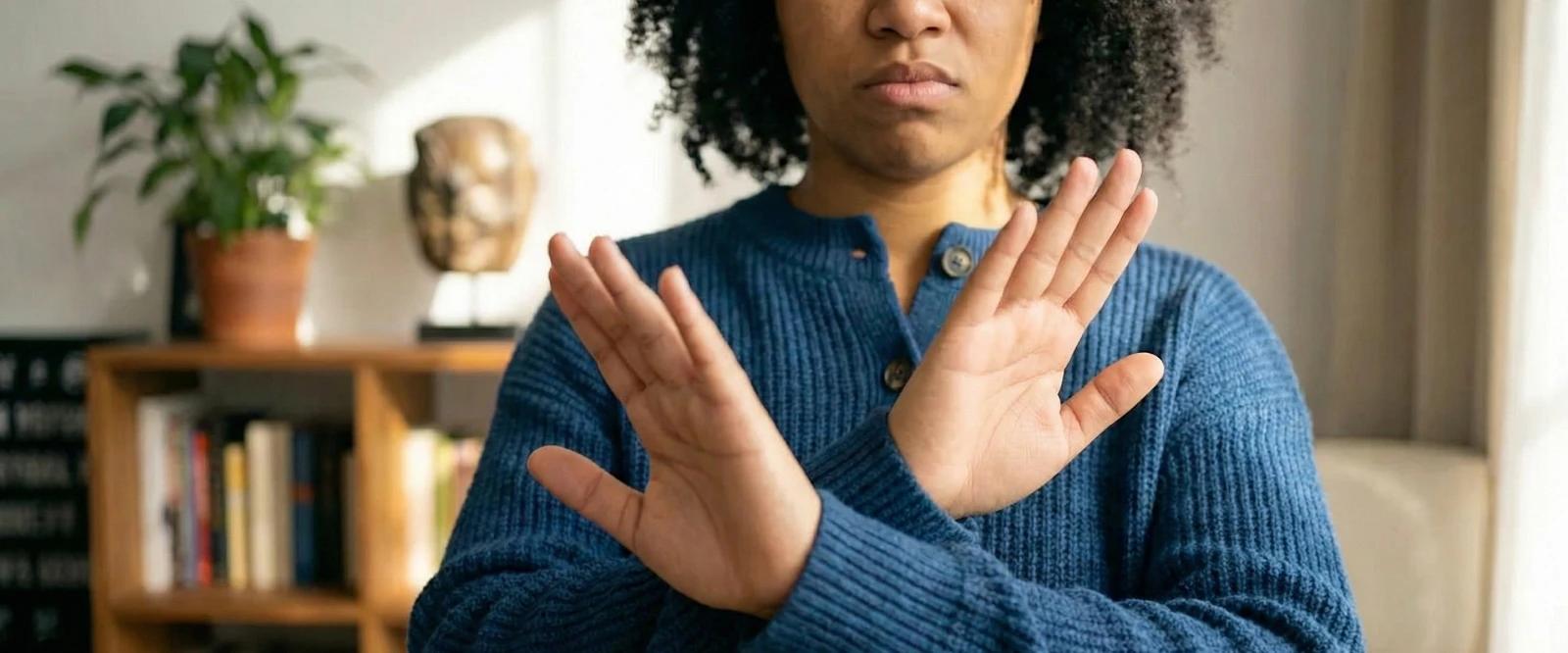 Black woman in a blue sweater crossing her arms in an X gesture, expressing refusal, with a bookshelf and potted plant in the background