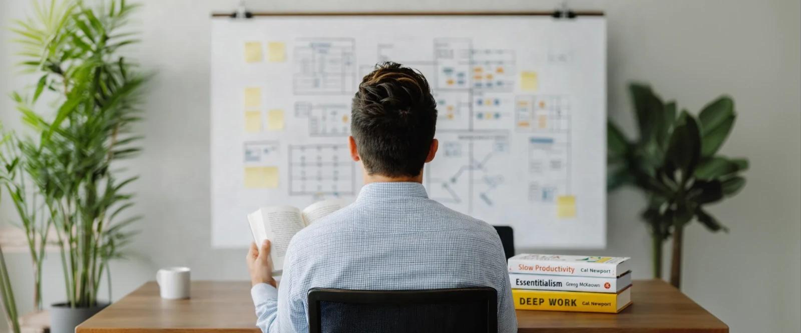 Person at desk with books for PMs including Deep Work, Essentialism, and Slow Productivity beside coffee mug, planning board, and green plants
