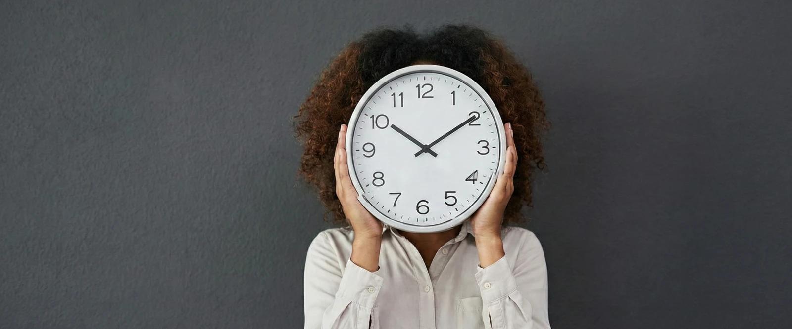 A woman with curly hair holding a large white clock in front of her face against a dark gray background, symbolizing time management and productivity