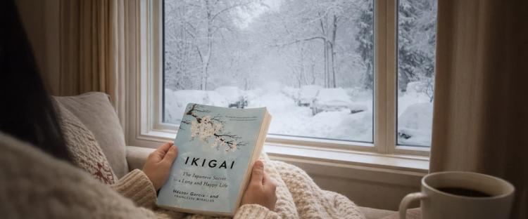 Hands holding Ikigai book by window with snow-covered trees outside, cozy setting for book summaries for a snowy weekend