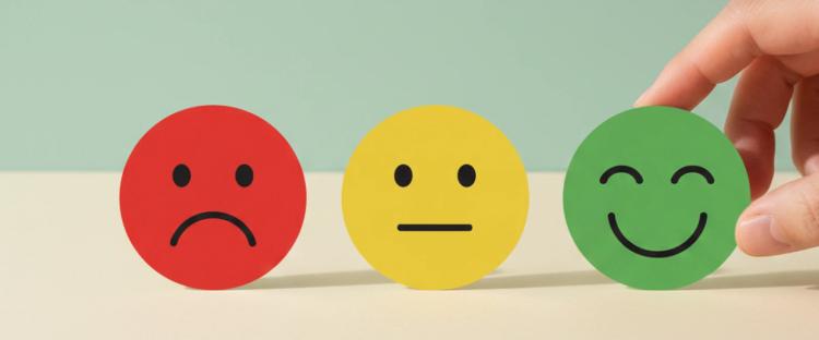 A hand picking the green happy emoji disc, next to yellow neutral and red sad emoji discs on a beige surface, against a soft teal background, symbolizing positive emotion and the law of attraction
