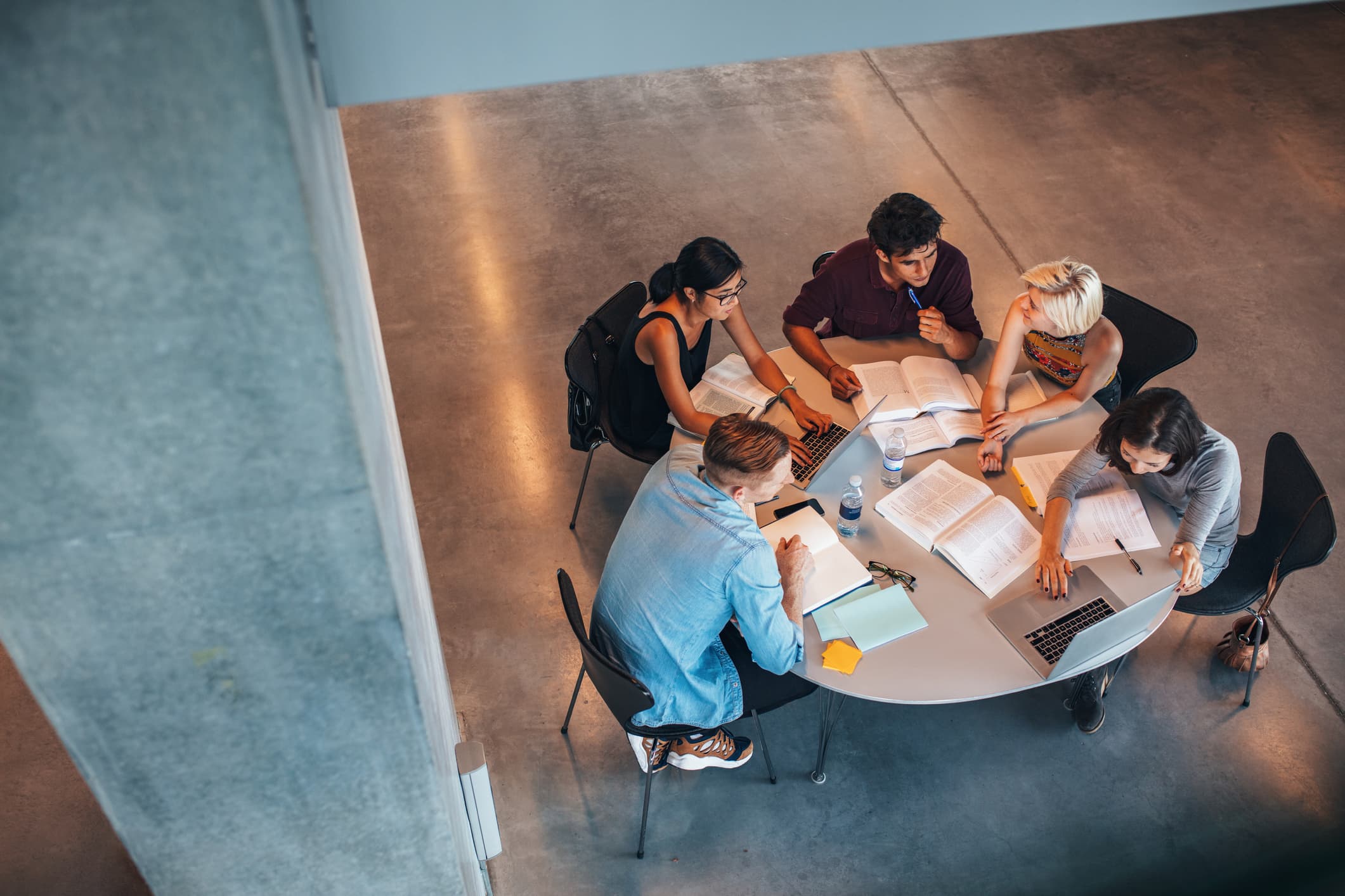 A group of young people sit around a table reading and talking together.