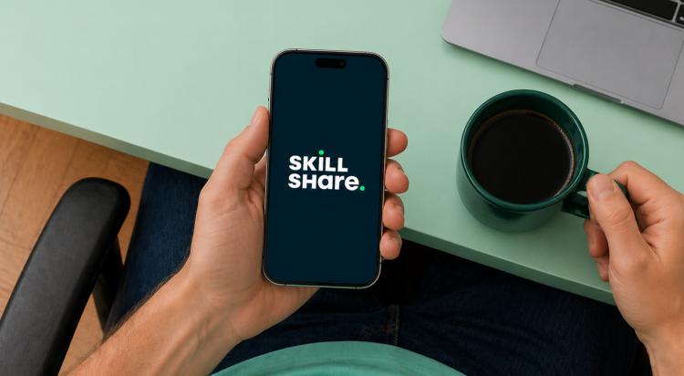 Person holding a smartphone with the Skillshare app logo on screen, green surface with a coffee cup and laptop in the background — Skillshare app review