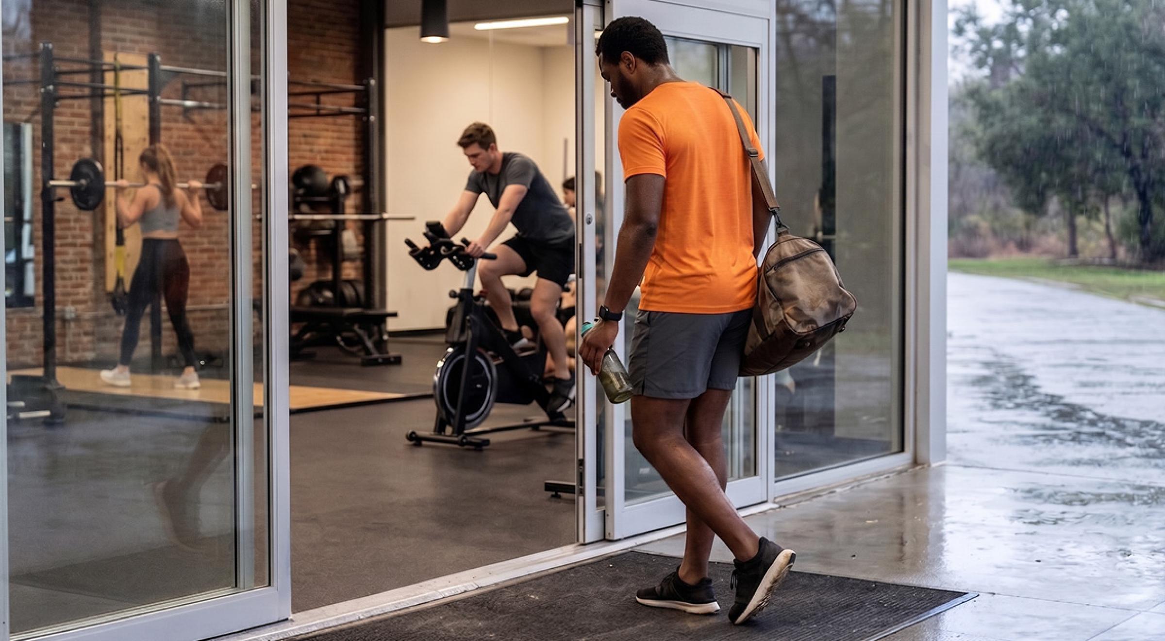 Man in orange t-shirt carrying a gym bag entering a fitness center in rainy weather, with people working out on exercise bike and barbell visible through glass doors inside