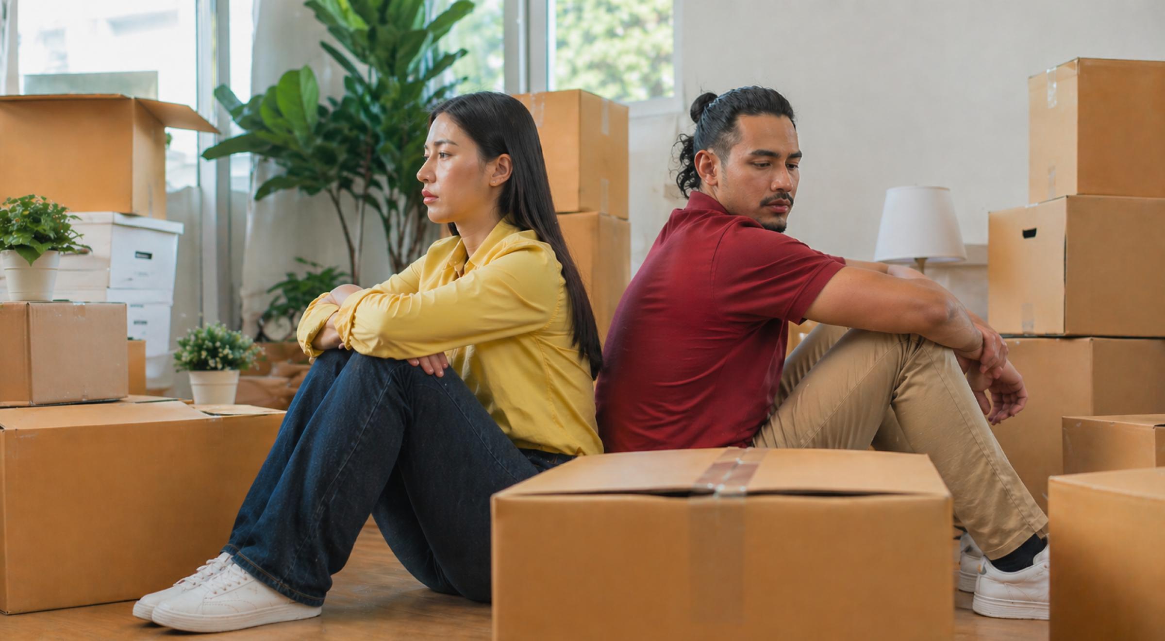 Couple sitting back-to-back on the floor surrounded by cardboard moving boxes, looking away from each other, depicting stages of a breakup in a relationship