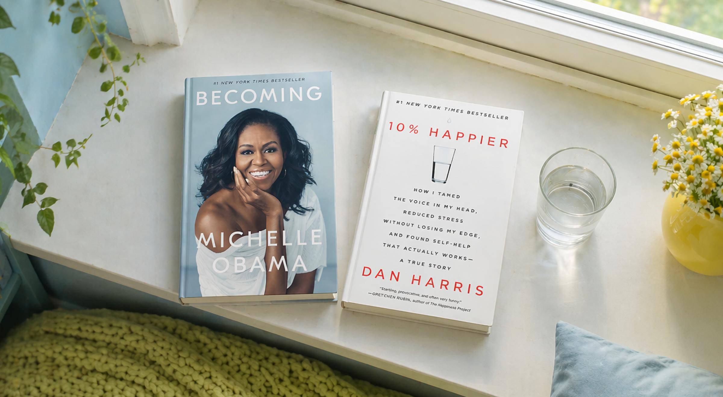 Becoming by Michelle Obama and 10% Happier by Dan Harris placed on a windowsill beside a glass of water and yellow flowers, illustrating memoir vs autobiography genres