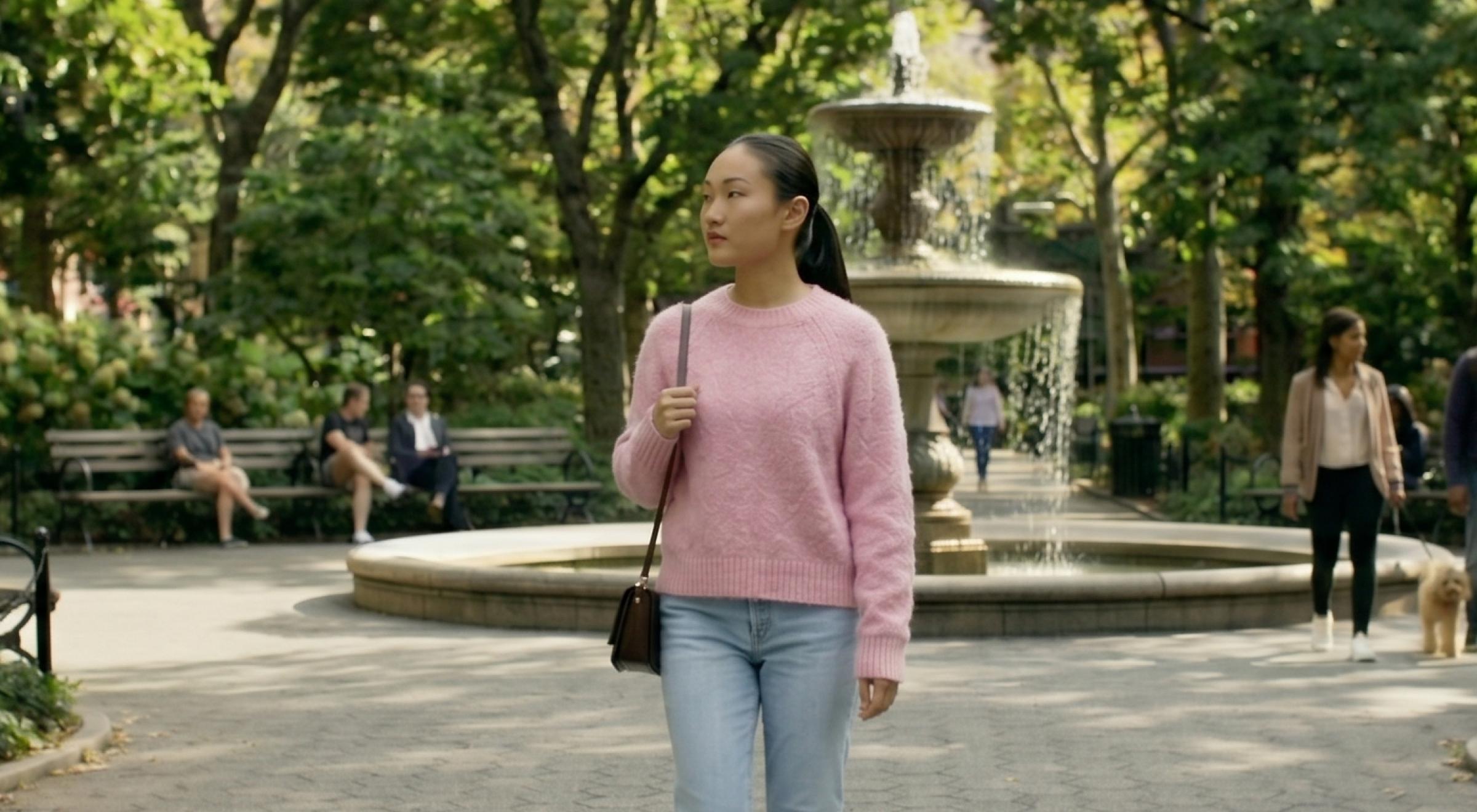 Young woman in a pink sweater and jeans standing alone near a city park fountain surrounded by green trees, reflecting on attachment after a break up