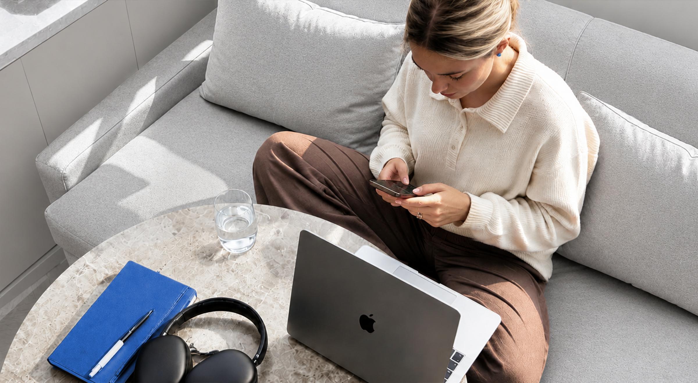 Young woman in a cream sweater sitting cross-legged on a grey sofa scrolling on her phone, with a MacBook, headphones, and blue notebook on a coffee table nearby
