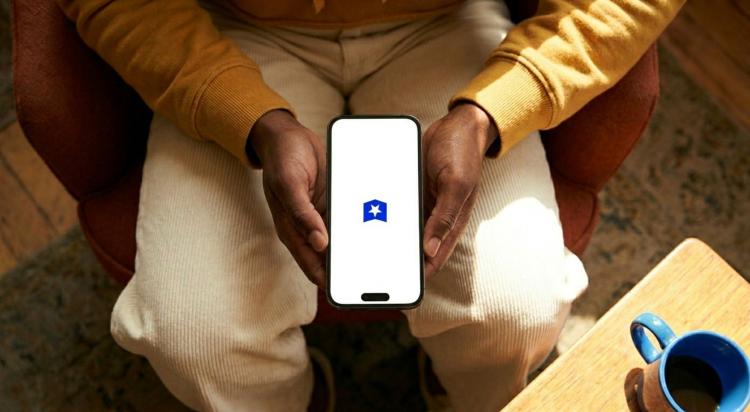 Person in a yellow sweater holding a smartphone with the Course Hero app logo while sitting in a cozy chair with a blue coffee mug nearby