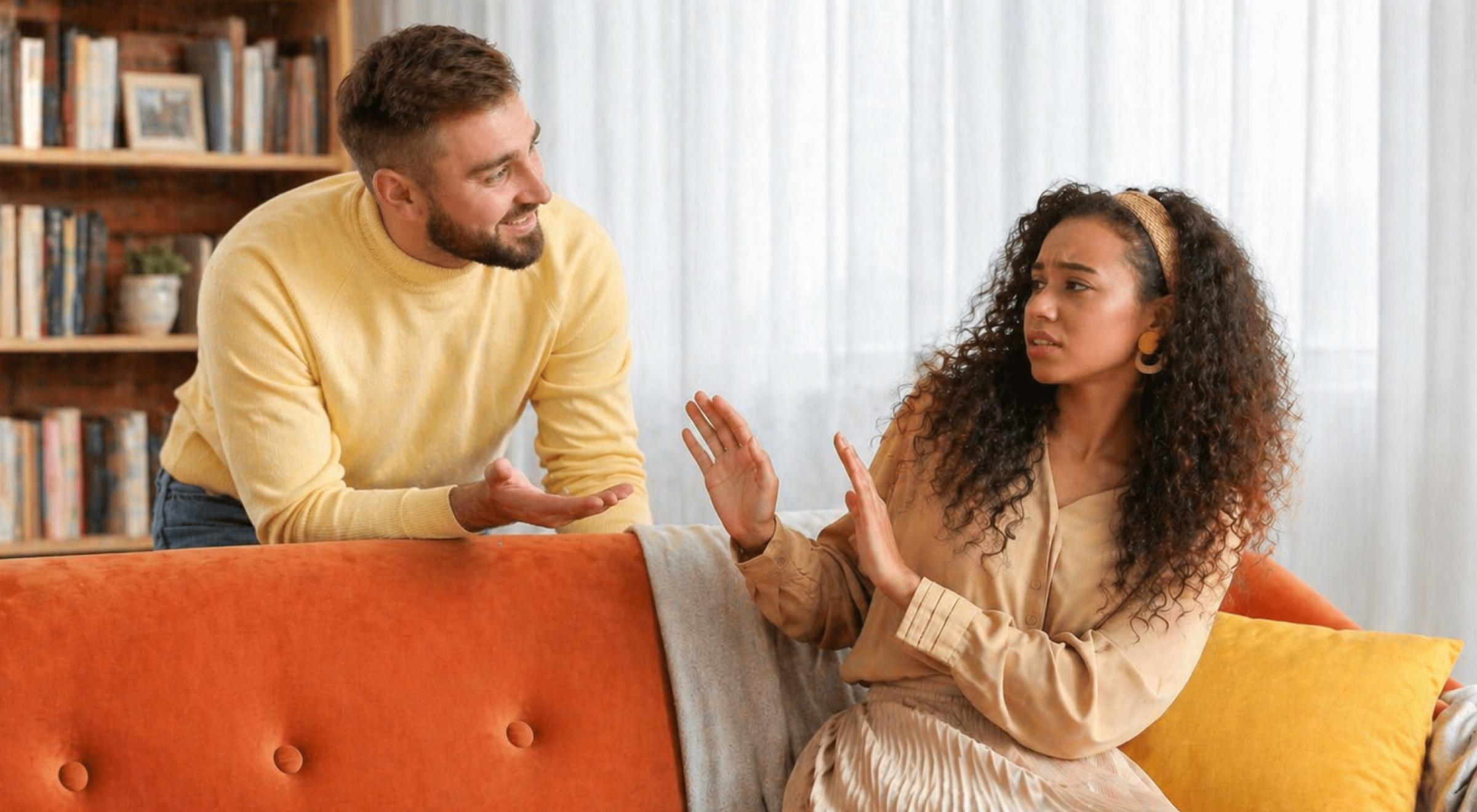 Man in yellow sweater and curly-haired woman arguing on an orange sofa, woman raising hand in rejection gesture, bookshelves visible in background — rejection skill in a relationship communication gui