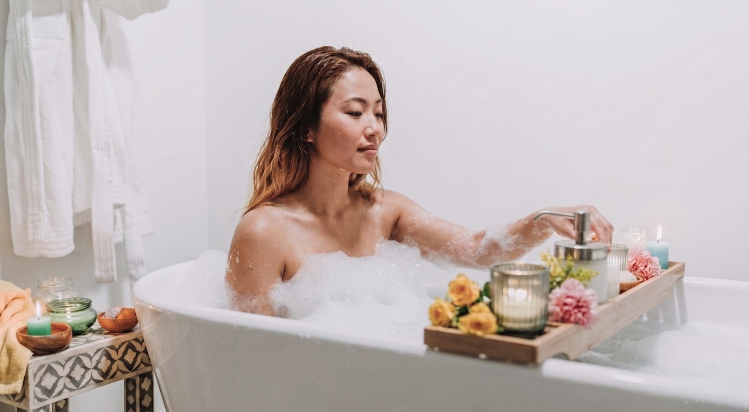 Young woman relaxing in a bubble bath with eyes closed, surrounded by candles and flowers on a wooden bath tray in a white bathroom