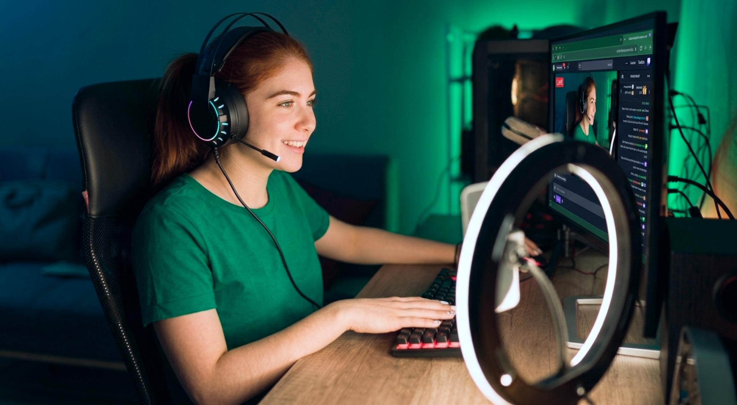 Young woman with headset and microphone streaming at a gaming desk with ring light and dual monitors glowing teal in a dark room