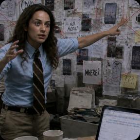 Woman in a blue shirt and tie gestures at a wall covered with notes and red string in a conspiracy investigation setup, with a laptop and coffee cups on the desk in front of her