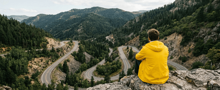 Man in yellow hoodie sits on cliff overlooking winding mountain road, symbolizing long path and determination required to make real change