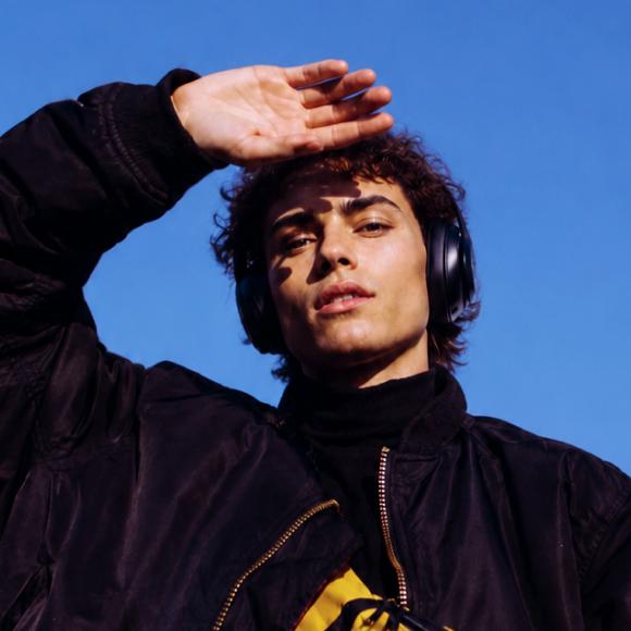 Young man wearing black over-ear headphones and a black bomber jacket, shielding his eyes with his hand against a clear blue sky while listening to the Headway app