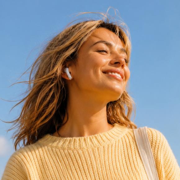 Smiling woman with wavy blonde hair wearing wireless earbuds and a yellow knit sweater outdoors against a clear blue sky, enjoying audio content from the Headway app