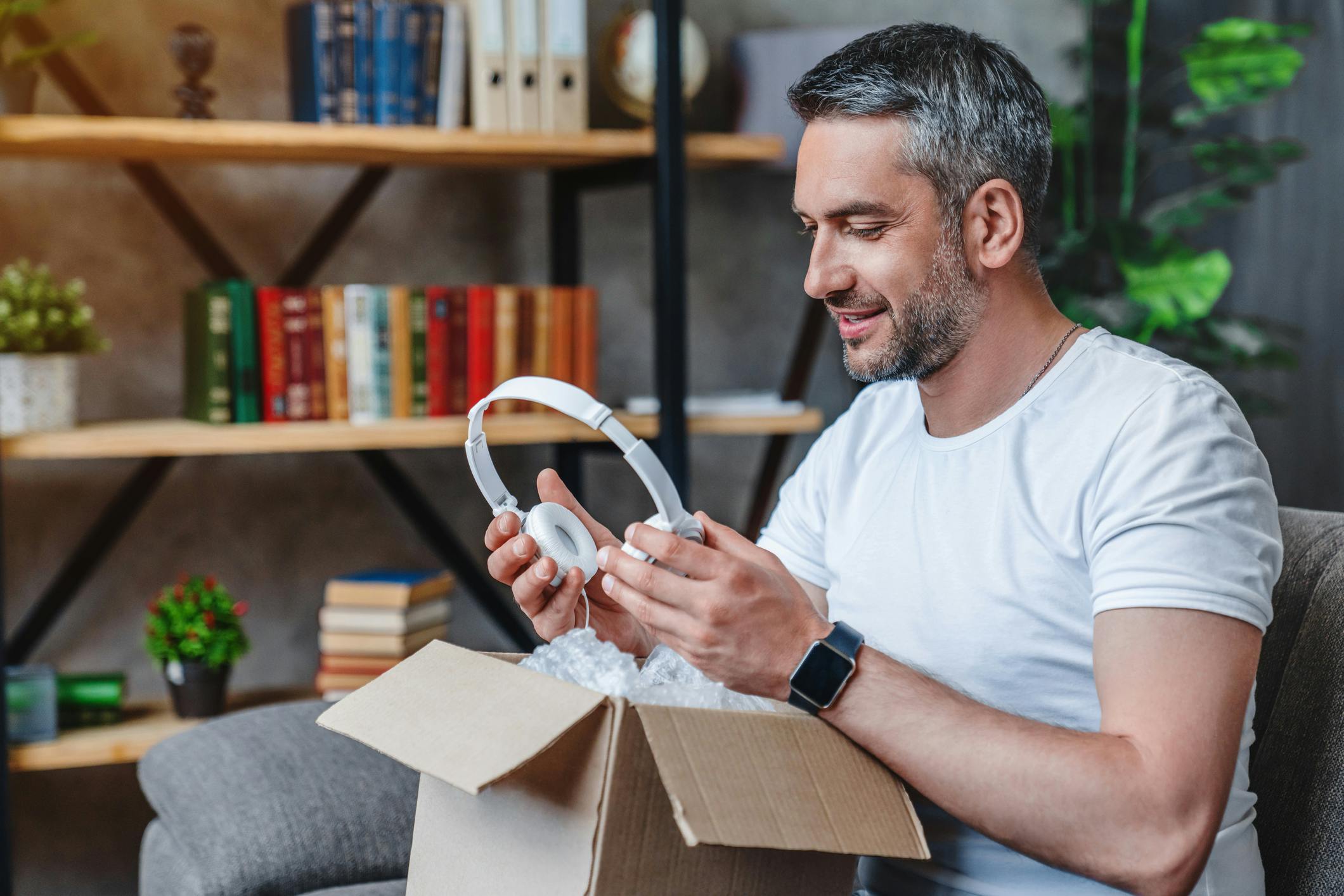 Next to a bookshelf, a man is opening a gift containing headphones.